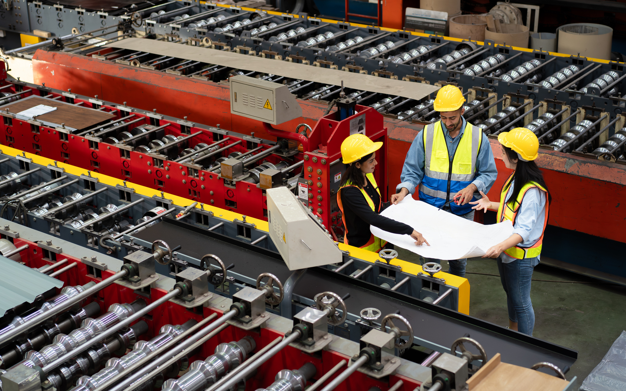 three people in safety gear stand beside a large metal forming machine reviewing a blueprint together in an active manufacturing environment