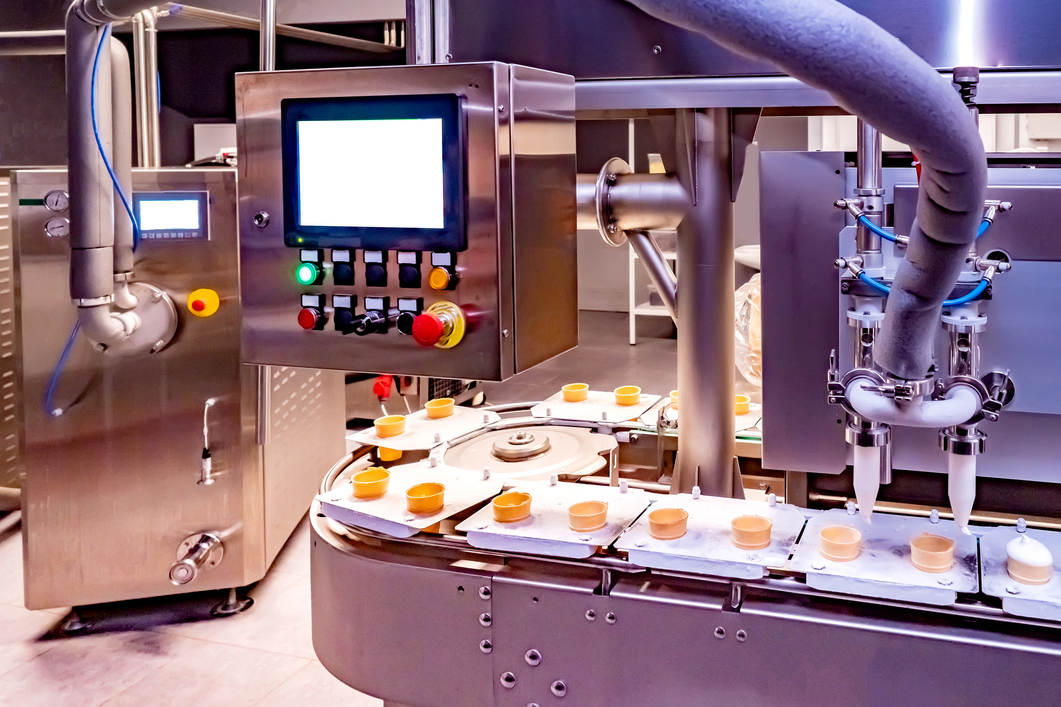 automated food‑processing equipment dispenses ice cream into containers on a conveyor line, with a control panel and stainless‑steel machinery visible in a clean production environment.