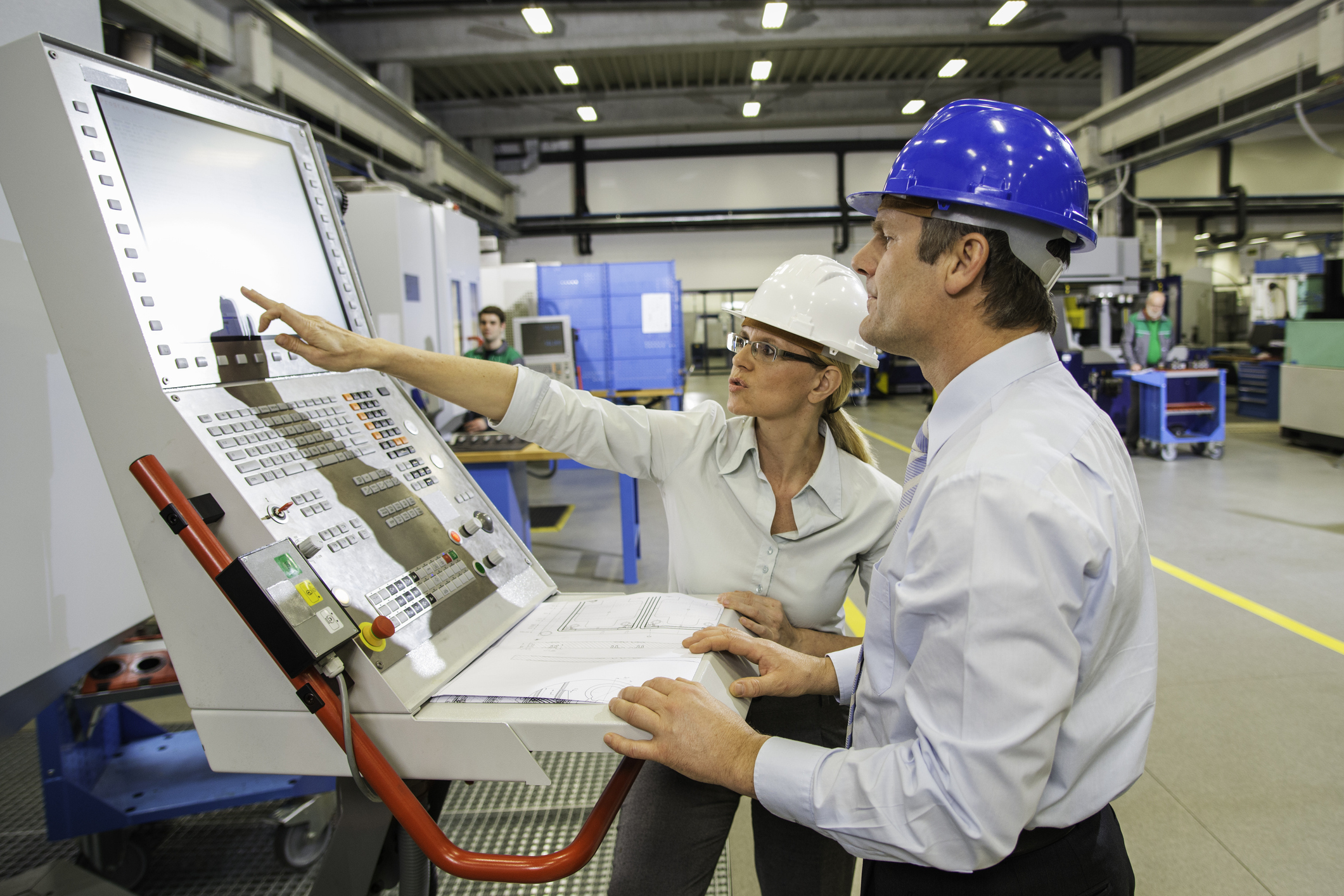 a pair of operators wearing safety helmets standing at a cnc control panel reviewing machine settings on the screen with printed schematics nearby illustrating how people interact directly with equipment through hmi style controls