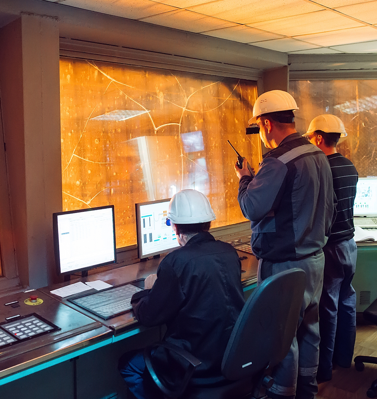 control room operators monitoring live steel production data with multiple screens and high‑heat processes visible through the windows illustrating real time analysis used to predict faults and maintain stable operations